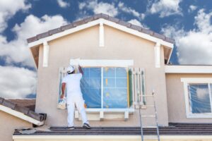 Painter working on the exterior of a house, painting the window frame with a roller brush on a sunny day, preparing the home for a fresh coat of paint. Ladder positioned nearby, with windows protected by plastic covering.